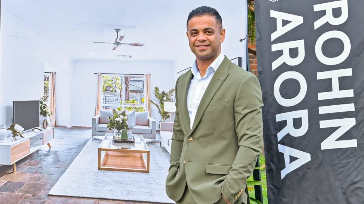 Rohin Arora in an olive blazer beside a Rohin Arora flag, in front of a styled living room