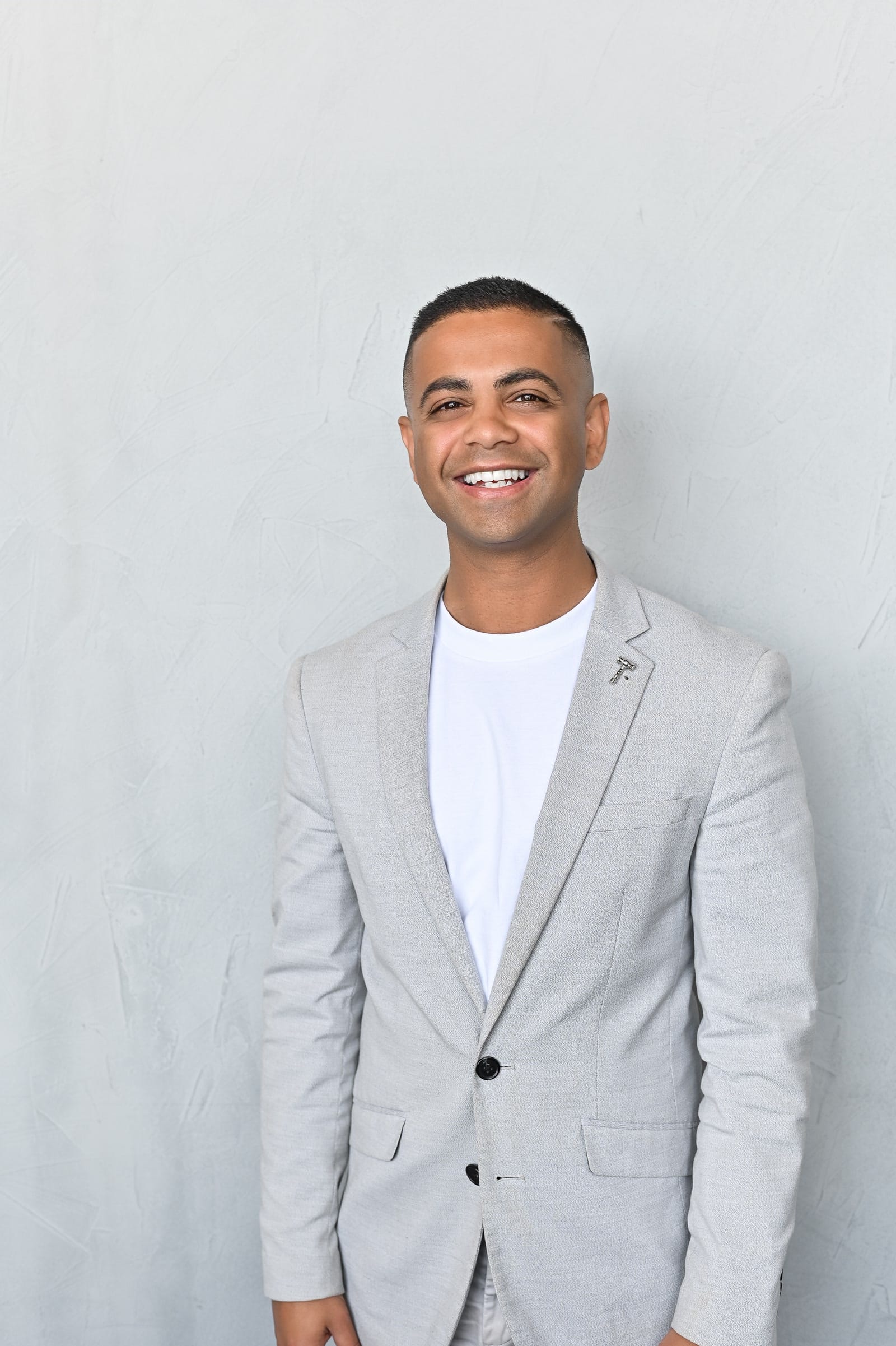Rohin Arora smiling, wearing a light grey blazer over a white t-shirt, standing against a pale plaster wall